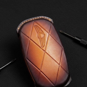 May include: A brown leather key fob with a diamond pattern and a gold emblem. The key fob is a warm brown color with a textured surface. Two small black screwdrivers are in the background.