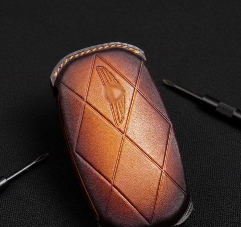 May include: A brown leather key fob with a diamond pattern and a gold emblem. The key fob is a warm brown color with a textured surface. Two small black screwdrivers are in the background.