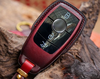 May include: Burgundy leather key fob cover with a black control panel and silver button. The cover has red stitching and a matching leather strap with a gold-colored metal clasp. The key fob is resting on a piece of wood.
