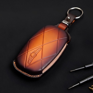 May include: A brown leather key fob case with a diamond pattern and a logo. The case has a metal ring and a leather strap. The case is approximately 4 inches long.