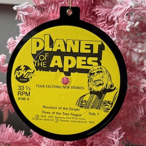 May include: A black and yellow Planet of the Apes Christmas ornament. The ornament is made from a record and features the movie title, a graphic of an ape, and text that reads "FOUR EXCITING NEW STORIES". The ornament hangs from a gold string.