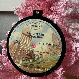 Könnte beinhalten: Ein Weihnachtsornament in Form einer Schallplatte des Albums Pyromania von Def Leppard. Der schwarze Schmuck hat eine goldene Schnur zum Aufhängen und zeigt das Albumcover und die Titelliste. Der Schmuck hängt an einem rosa Weihnachtsbaum.