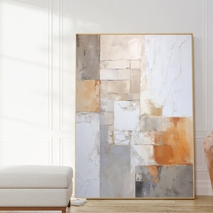 May include: Abstract painting with a geometric design in shades of beige, brown, and white. The painting is framed in a thin gold frame and is hanging on a white wall.
