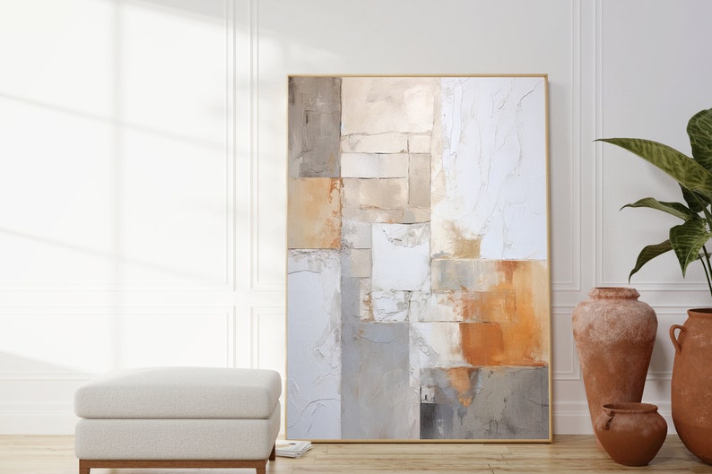 May include: Abstract painting with a geometric design in shades of beige, brown, and white. The painting is framed in a thin gold frame and is hanging on a white wall.