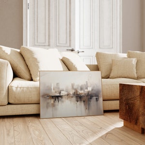 May include: A framed abstract painting of a city skyline with a reflection in the water. The painting is in shades of gray, brown, and white. It is hanging on a wall above a beige couch.