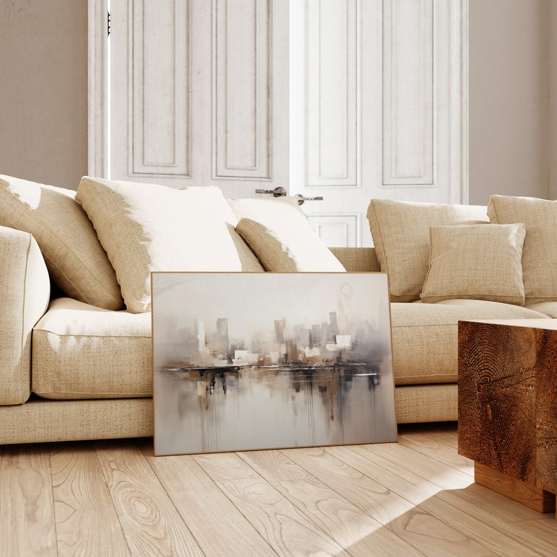 May include: A framed abstract painting of a city skyline with a reflection in the water. The painting is in shades of gray, brown, and white. It is hanging on a wall above a beige couch.