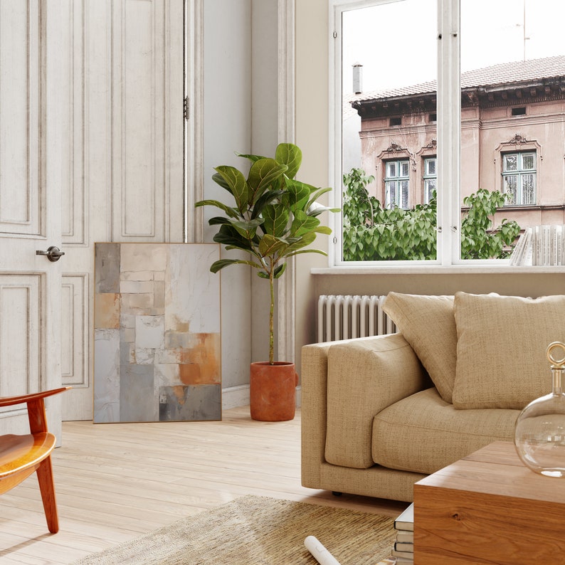 May include: A living room interior with a beige sofa, a potted ficus plant, and a framed abstract painting on the wall. The sofa is positioned in front of a window with a view of a city street.