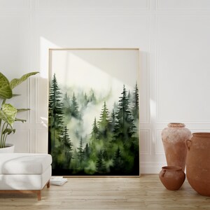 Op de afbeelding: Een aquarel schilderij van een bos sc&egrave;ne met hoge groene bomen en een mistige achtergrond. Het schilderij is ingelijst in een gouden lijst en hangt aan een witte muur.
