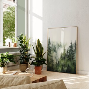 Op de afbeelding: Een ingelijst aquarel schilderij van een mistige bossen sc&egrave;ne met hoge groene bomen. Het schilderij hangt aan een witte muur in een kamer met planten en een houten salontafel.