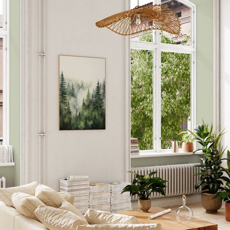 Op de afbeelding: Een woonkamerinterieur met een groot raam, een beige bank en een ingelijst aquarel schilderij van een bos sc&egrave;ne. Het schilderij hangt aan een witte muur. Er staat een grote potplant in de hoek van de kamer.