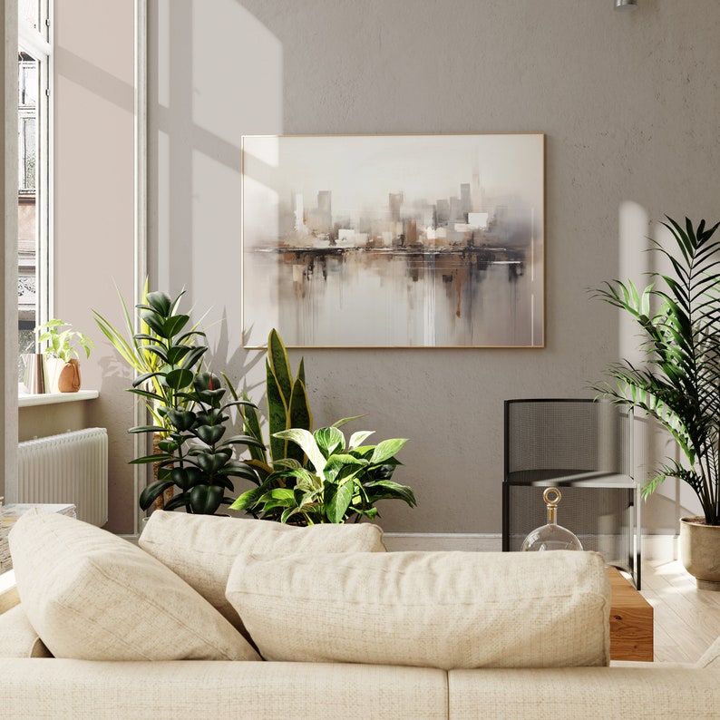May include: A beige couch with three pillows in a living room with a large abstract painting of a city skyline in muted brown and beige tones. There are several green plants in the room, including a snake plant and a ficus.