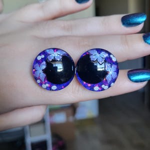 May include: A pair of round earrings with a black center and a purple and white floral design. The earrings have a glossy finish and are set against a blurred background.
