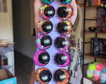 May include: A set of fourteen round cabochon craft eyes arranged in two vertical columns on a light purple card. Each eye features a large black pupil surrounded by a vibrant, multi-colored glitter iris in shades of green, pink, orange, and gold.