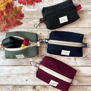 May include: Four small, zippered pouches in shades of black, green, navy, and burgundy. Each pouch has a contrasting zipper and a small fabric tag that reads "Sweet W". The pouches are rectangular with a small loop and a metal clasp.