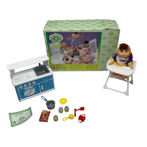 May include: A Cabbage Patch Kids Feeding Playset with a blue and white stove, a white high chair, and a doll with a red and yellow outfit. The doll is sitting in the high chair and has a red bow in her hair. The set also includes a variety of accessories, such as a pot, a spoon, and a bottle.