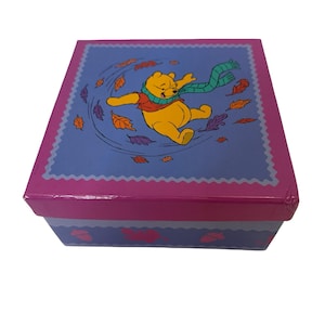 May include: A square box with a lid featuring a cartoon illustration of Winnie the Pooh. The lid is blue with a pink border and a repeating leaf pattern. Winnie the Pooh is wearing a scarf and surrounded by falling leaves.