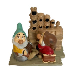 May include: Two dwarf figurines from the Disney movie Snow White and the Seven Dwarfs. The dwarfs are playing music on a wooden organ with carved faces. The organ is brown and the dwarfs are wearing colorful clothing.