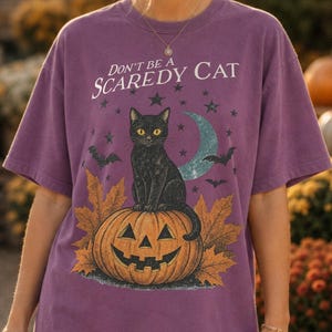 Pode incluir: Uma t-shirt roxa de manga curta com uma estampa gráfica de um gato preto sentado numa abóbora de Halloween rodeado por folhas de outono, estrelas e uma lua crescente, com o texto Don't be a SCAREDY CAT impresso acima da ilustração.