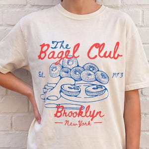 Può includere: T-shirt color crema con la scritta "The Bagel Club Brooklyn New York" in rosso e blu, con un disegno di bagel. La maglietta a maniche corte è indossata da una persona. Il testo include l'anno 1973.