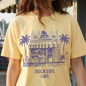 Peut inclure: T-shirt jaune clair avec un motif violet d'un caf&eacute;, de palmiers et le texte "DOCKSIDE CAFE". La personne porte un short en jean et tient une boisson. L'arri&egrave;re-plan comprend un b&acirc;timent, des bateaux et des fleurs.