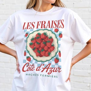 May include: White t-shirt featuring a strawberry graphic with the text "LES FRAISES" and "Cote d'Azur". The design uses red and green, with a blue and white checkered pattern. The shirt is a casual style.