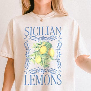 A woman is wearing an Ivory Comfort Colors shirt with a design featuring a branch of lemons and the phrase Sicilain Lemons.