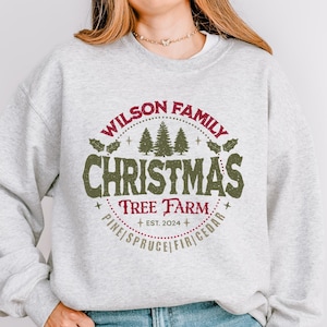 Pode incluir: Uma camisa de frio cinza com um design de fazenda de árvores de Natal vermelho e verde. O design apresenta o texto "Wilson Family Christmas Tree Farm + EST. 2024 + PINE SPRUCE/FIR CEDAR".