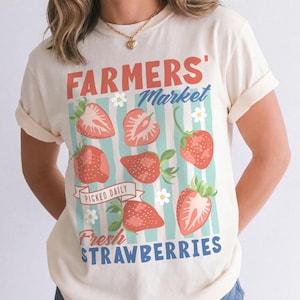 May include: Cream-colored t-shirt with a "FARMERS' MARKET" graphic featuring strawberries and the text "PICKED DAILY FRESH STRAWBERRIES". The design includes red strawberries, blue stripes, and white flowers. The shirt has rolled-up sleeves.