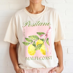 A woman is wearing a Natural Gildan 2000 unisex T-shirt with a design featuring a branch of lemons with a long pink coquette bow tied around it and the phrase Positano, Amalfi Coast, Italy.