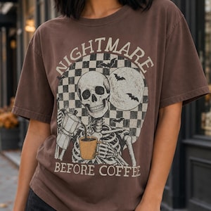 Op de afbeelding: Een bruin t-shirt met korte mouwen en een afbeelding van een skelet dat koffie uit een mokkapot in een mok schenkt. Het ontwerp bevat een geblokte achtergrond, vleermuizen, een maan en de tekst "NIGHTMARE BEFORE COFFEE" in witte letters.