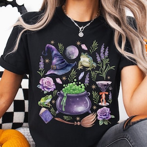 May include: Black t-shirt with a witchy design featuring a witch's hat, cauldron, frogs, roses, a potion bottle, a crystal, a moon, and a broom.