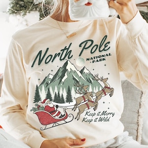 May include: Ivory long-sleeve shirt with a graphic of Santa Claus in a sleigh, mountains, and the text "North Pole National Park." The shirt also has the text "Keep it Merry, Keep it Wild." A Santa mug is held up.
