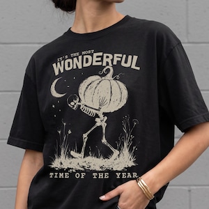 Puede incluir: Una camiseta negra de manga corta con un gráfico en color beige de un esqueleto cargando una calabaza grande. El texto dice: "IT'S THE MOST WONDERFUL TIME OF THE YEAR". El diseño incluye una luna creciente, estrellas y hierba alta en la ilustración.