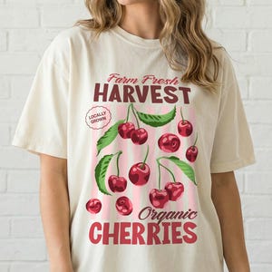 May include: A cream-coloured t-shirt with a graphic of cherries and green leaves, and the text "Farm Fresh Harvest Organic Cherries." The design includes pink vertical stripes and the words "Locally Grown."