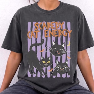 Pode incluir: Uma t-shirt de manga curta cinzento-carvão com o texto laranja "SCAREDY CAT ENERGY" sobre um gráfico de três gatos pretos, pequenos morcegos cor de laranja e uma lua crescente amarela sobre um fundo de riscas verticais roxas.