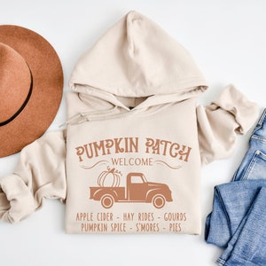 Peut inclure: Un sweat à capuche beige avec un graphique brun d'un camion transportant une citrouille. Le graphique dit "Pumpkin Patch Welcome" et "Apple Cider - Hay Rides - Gourds - Pumpkin Spice - S'mores - Pies".