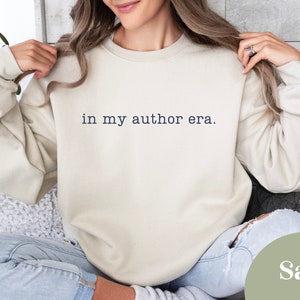 May include: A sand-coloured sweatshirt featuring the phrase "in my author era." This long-sleeved crew neck top is a neutral colour, suitable for casual wear. A comfortable and stylish choice for writers and authors.