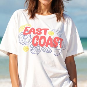 Puede incluir: Una camiseta blanca con un diseño gráfico de conchas y limones. El texto "East Coast" está impreso en rojo en la camiseta.