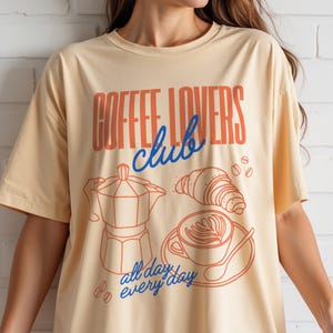 A woman is wearing a Vegas Gold Gildan 2000 t-shirt with a design featuring a hand-drawn coffee and croissant design that reads Coffee Lover Club, All Day Every Day.