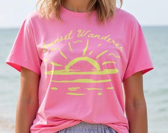 May include: A pink t-shirt with the text "Sunset Wanderer" in yellow, featuring a sun graphic. The shirt is paired with blue and white striped shorts. The image is taken on a beach with the ocean in the background.