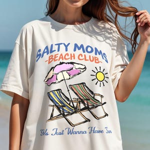 A woman is standing on a beach wearing an oversized Comfort Colors t-shirt with a design featuring a colorful hand-drawn scene of a striped beach umbrella and two chairs with the phrase Salty Moms Beach Club at the top and Girls Just Wanna Have Fun at the bottom of the design.