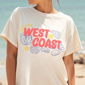 Puede incluir: Una camiseta blanca de gran tamaño con un estampado gráfico de las palabras "WEST COAST" en rosa y amarillo con ilustraciones azules y blancas de conchas y limones.