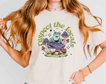 Op de afbeelding: Een licht beige t-shirt met een afbeelding van een groene kikker die op een paarse paddenstoel zit met de tekst "respect the locals" in groene en gele letters.