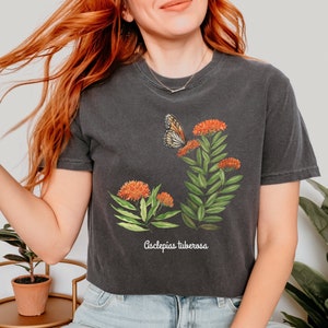 Op de afbeelding: Een donkergrijs gecropped t-shirt met een botanische illustratie van oranje bloemen en een vlinder. De tekst "Asclepias tuberosa" is onder de illustratie gedrukt.