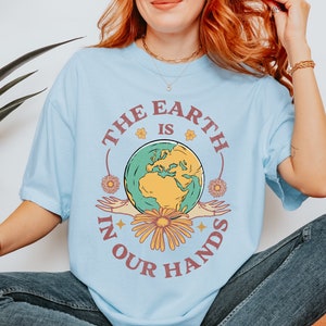 A woman is wearing a chambray blue Comfort Colors shirt with an Earth Day design that says The Earth is in Your Hands.