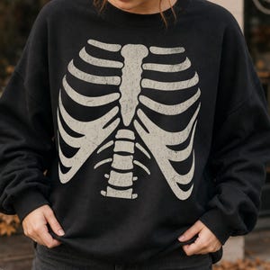 May include: A black crewneck sweatshirt featuring a large, centred, off-white graphic print of a human rib cage and sternum. The skeletal design has a distressed, weathered texture and covers the front torso area of the long-sleeved garment.