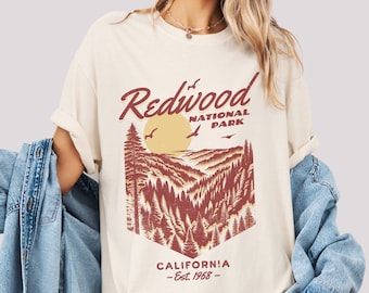 May include: Cream-colored t-shirt featuring a Redwood National Park graphic. The design includes the words "Redwood National Park" in a script font, a sun, birds, and a forest scene. The text "CALIFORNIA - Est. 1968 -" is also included.