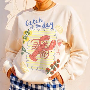 May include: Cream-colored sweatshirt with a lobster graphic and the text "Catch of the Day." The graphic includes a red lobster, a glass of wine, and lemon slices. The person is wearing blue and white checkered shorts.