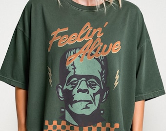 Puede incluir: Camiseta verde oscuro con un gráfico del monstruo de Frankenstein y el texto "Feelin' Alive" en naranja. El diseño incluye rayos y un patrón de cuadros. La camiseta tiene mangas cortas.