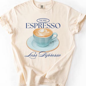 A flat lay of an Ivory Comfort Colors shirt with a design featuring an espresso and the phrase More Espresso Less Depresso.
